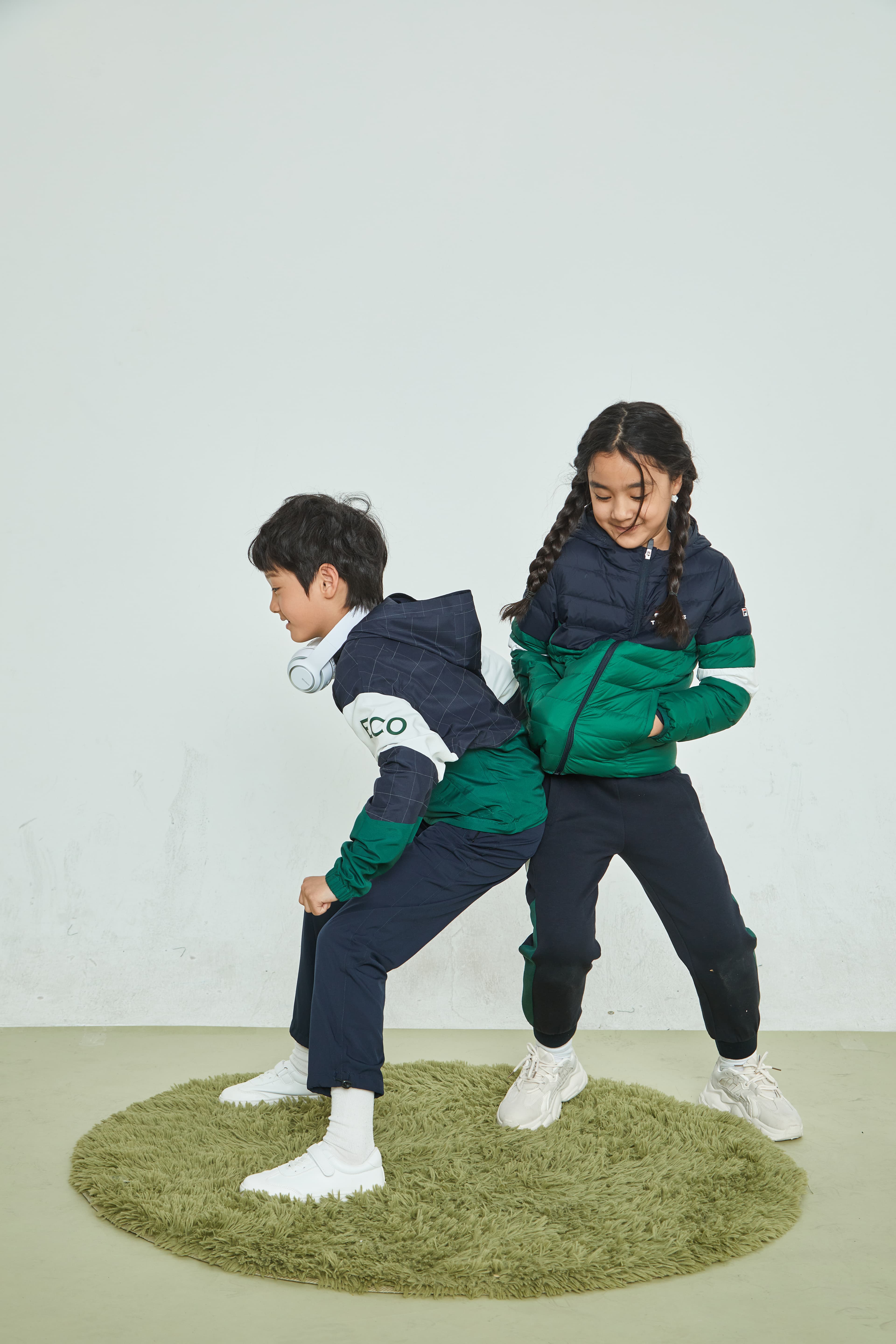 Two children in navy and green sporty outfits pose back-to-back on a green rug.