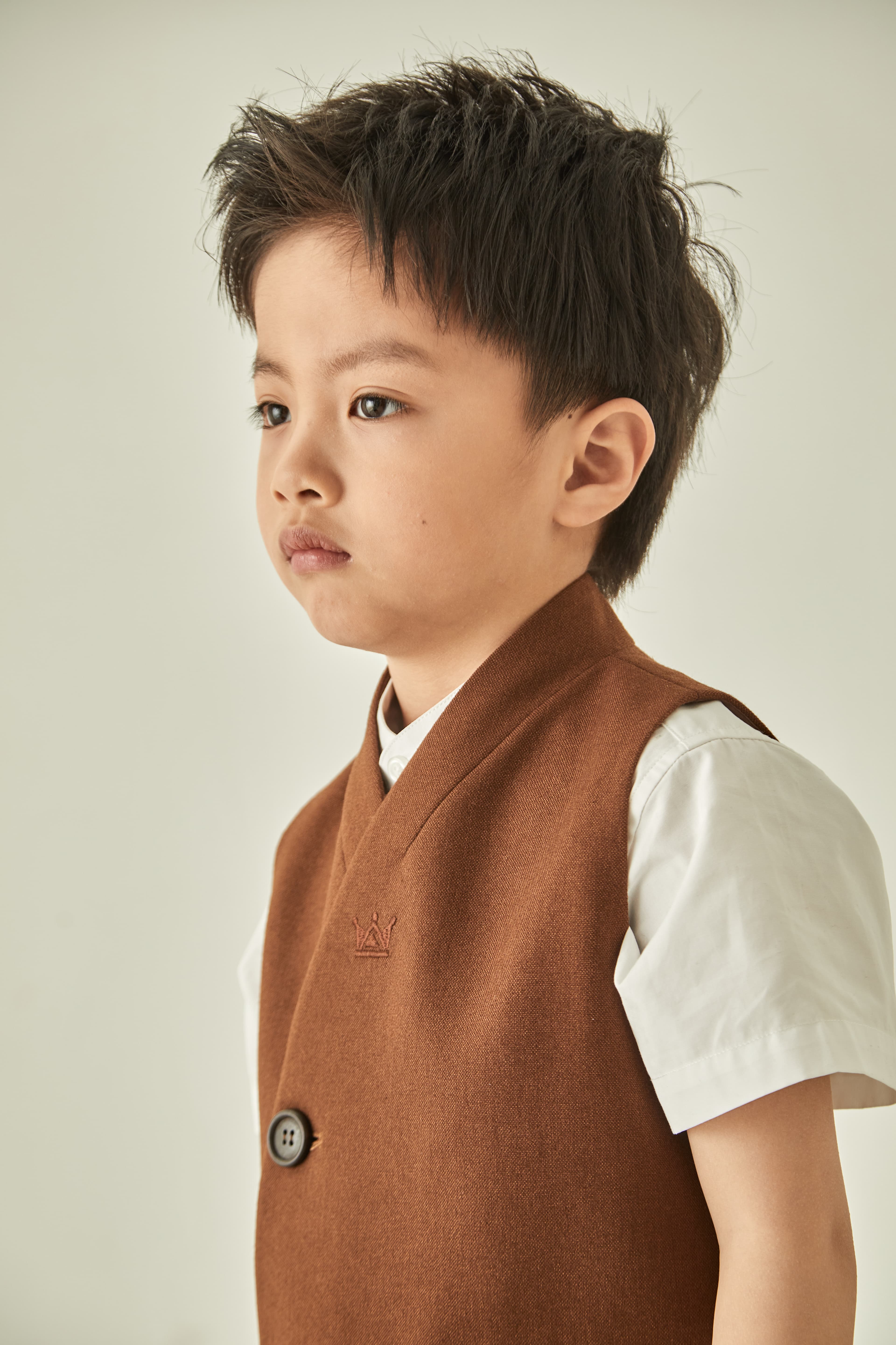 Young boy with messy dark hair looking away, wearing a brown vest and white shirt.