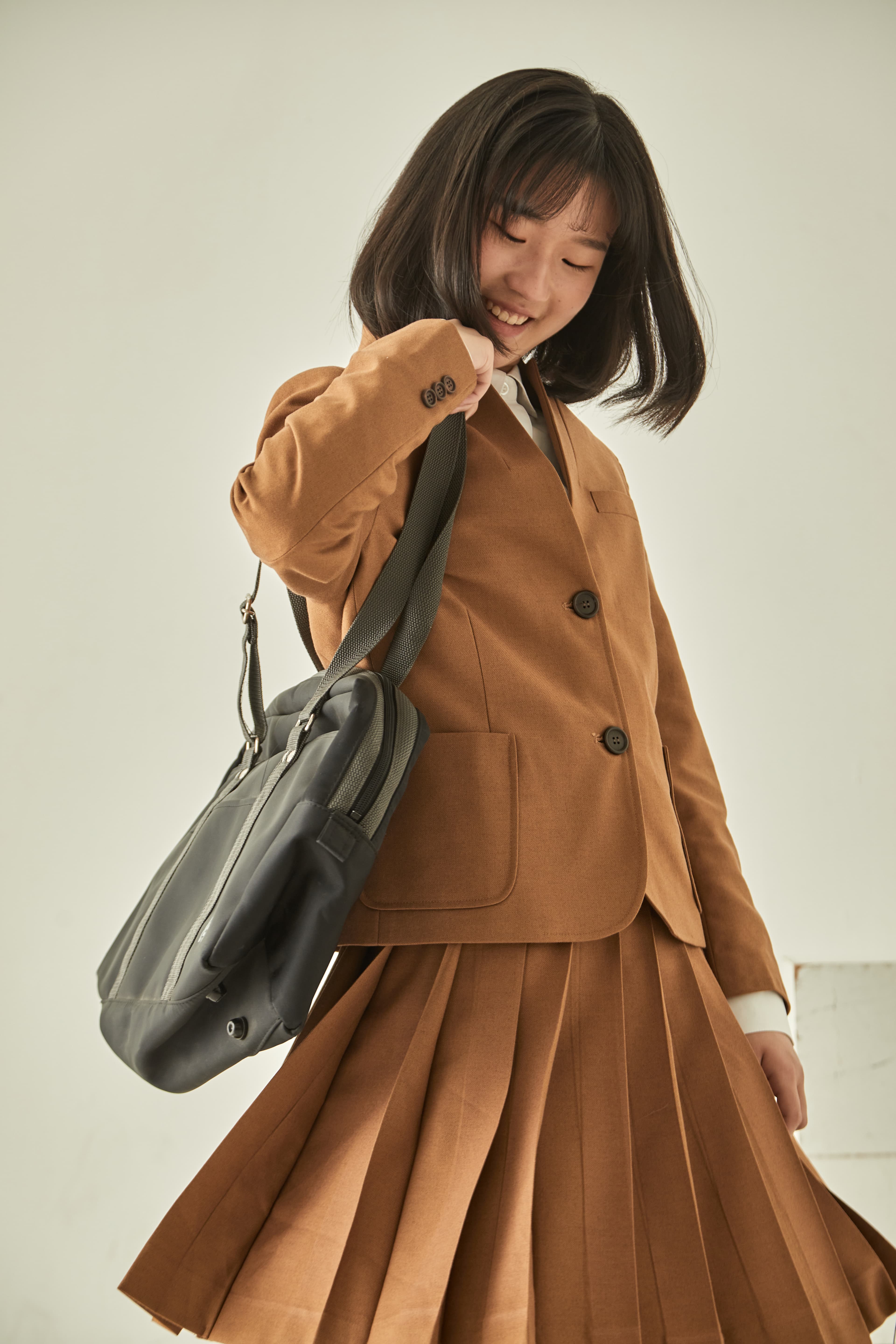 Smiling student in a brown blazer and pleated skirt carrying a black shoulder bag.