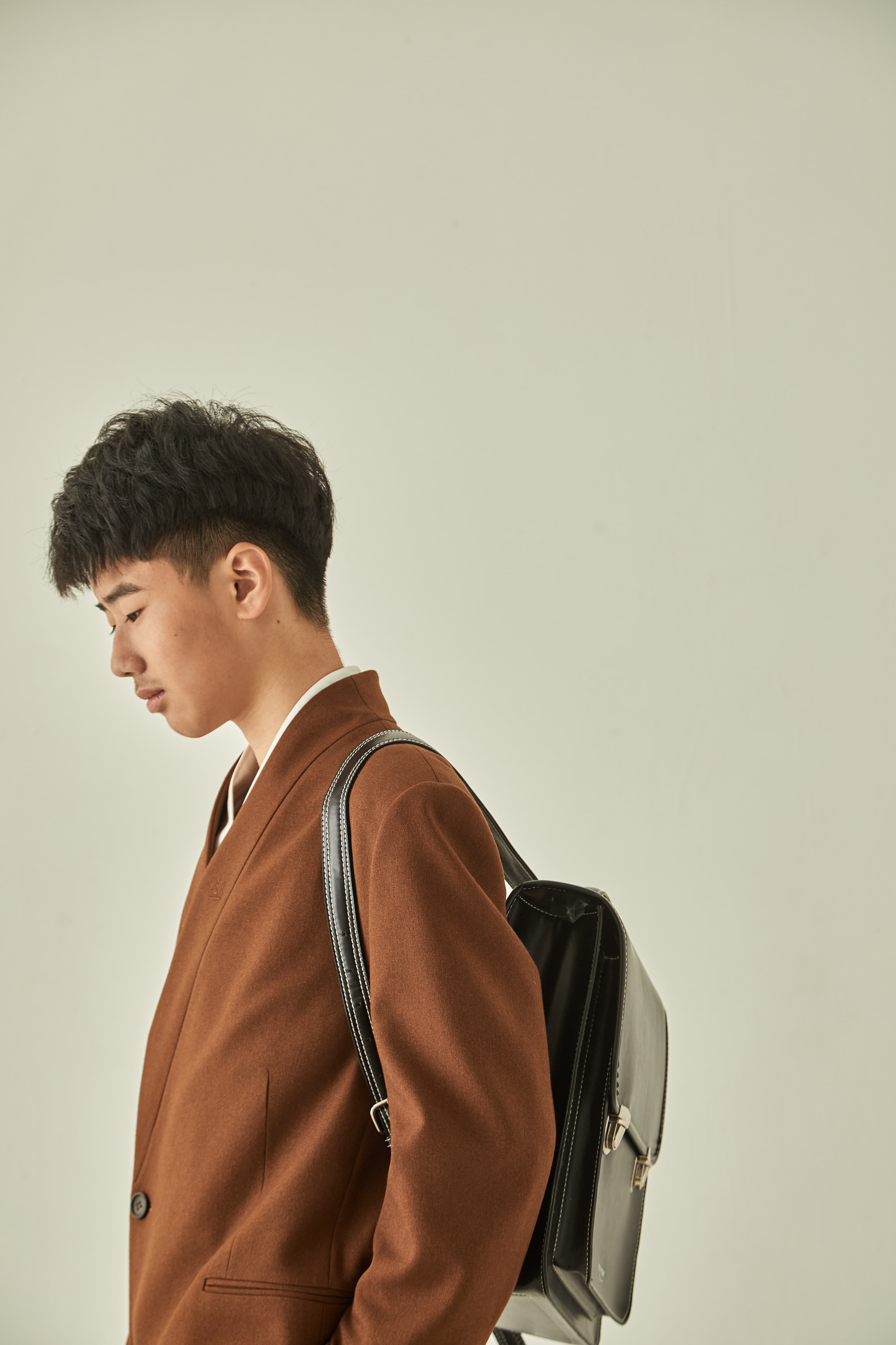 Young man in a brown blazer looking down while wearing a black leather backpack.
