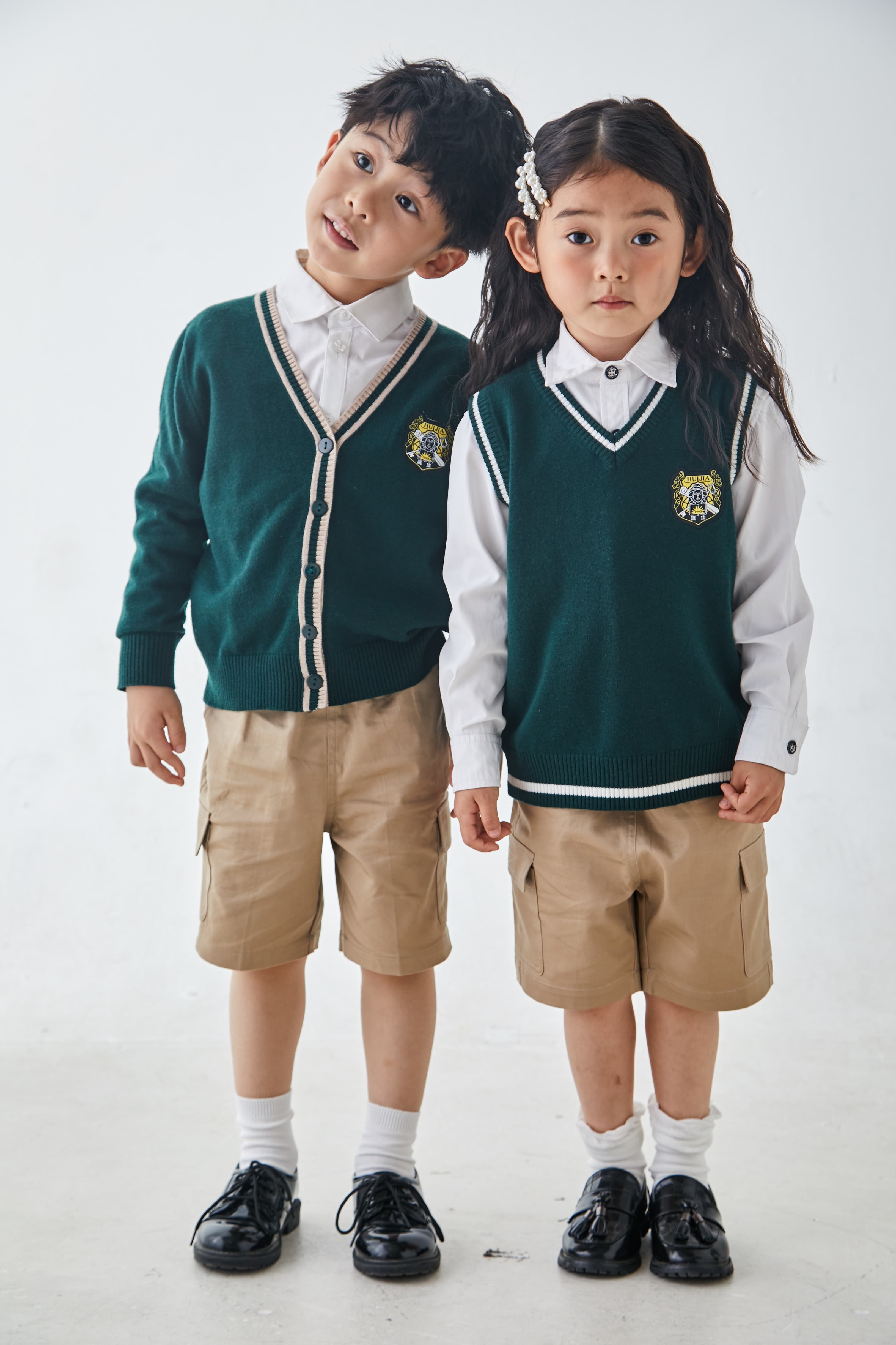 Two children wearing matching green school sweaters and tan shorts against a white background.