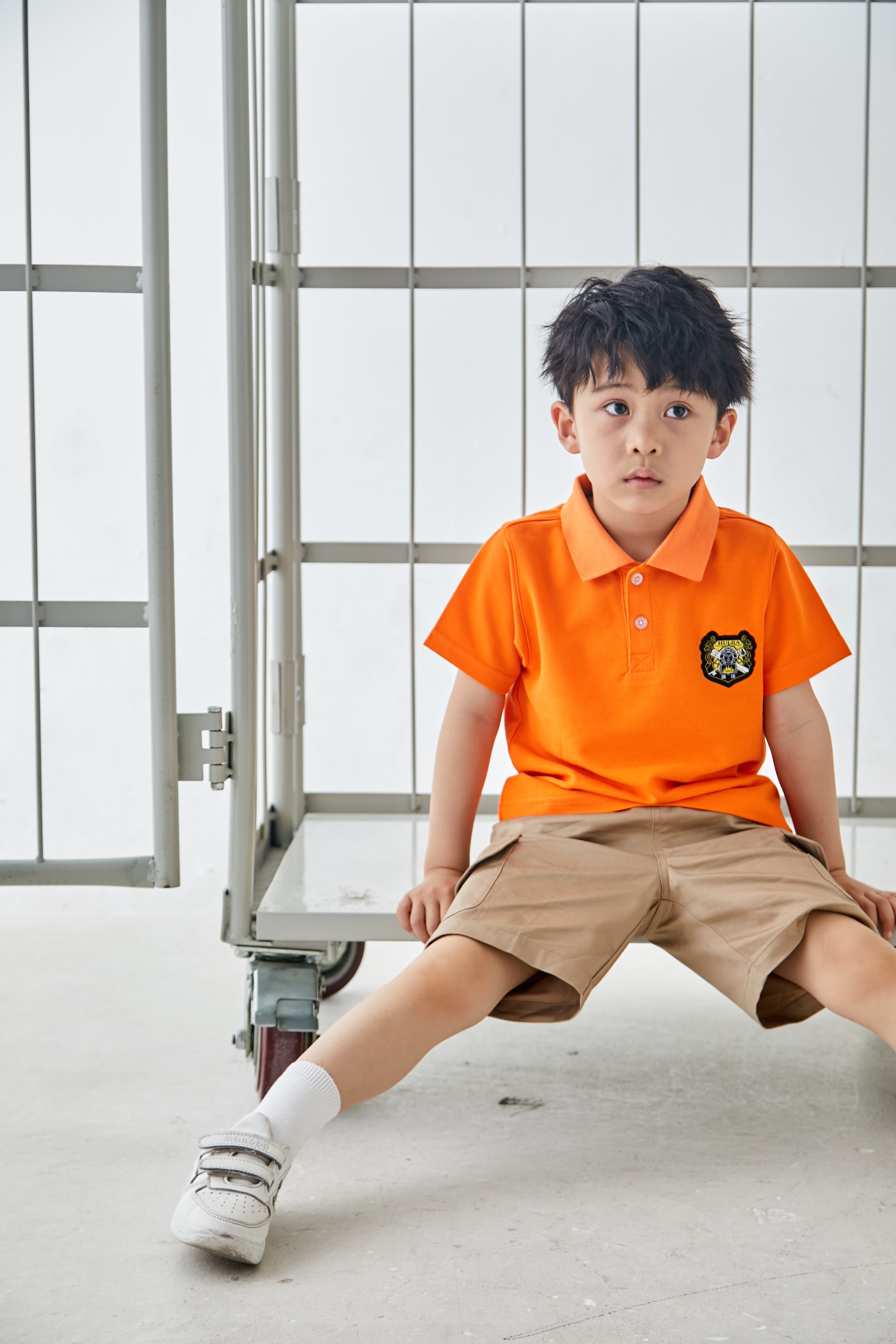 Young boy wearing an orange polo shirt and khaki shorts sitting on a metal cart.