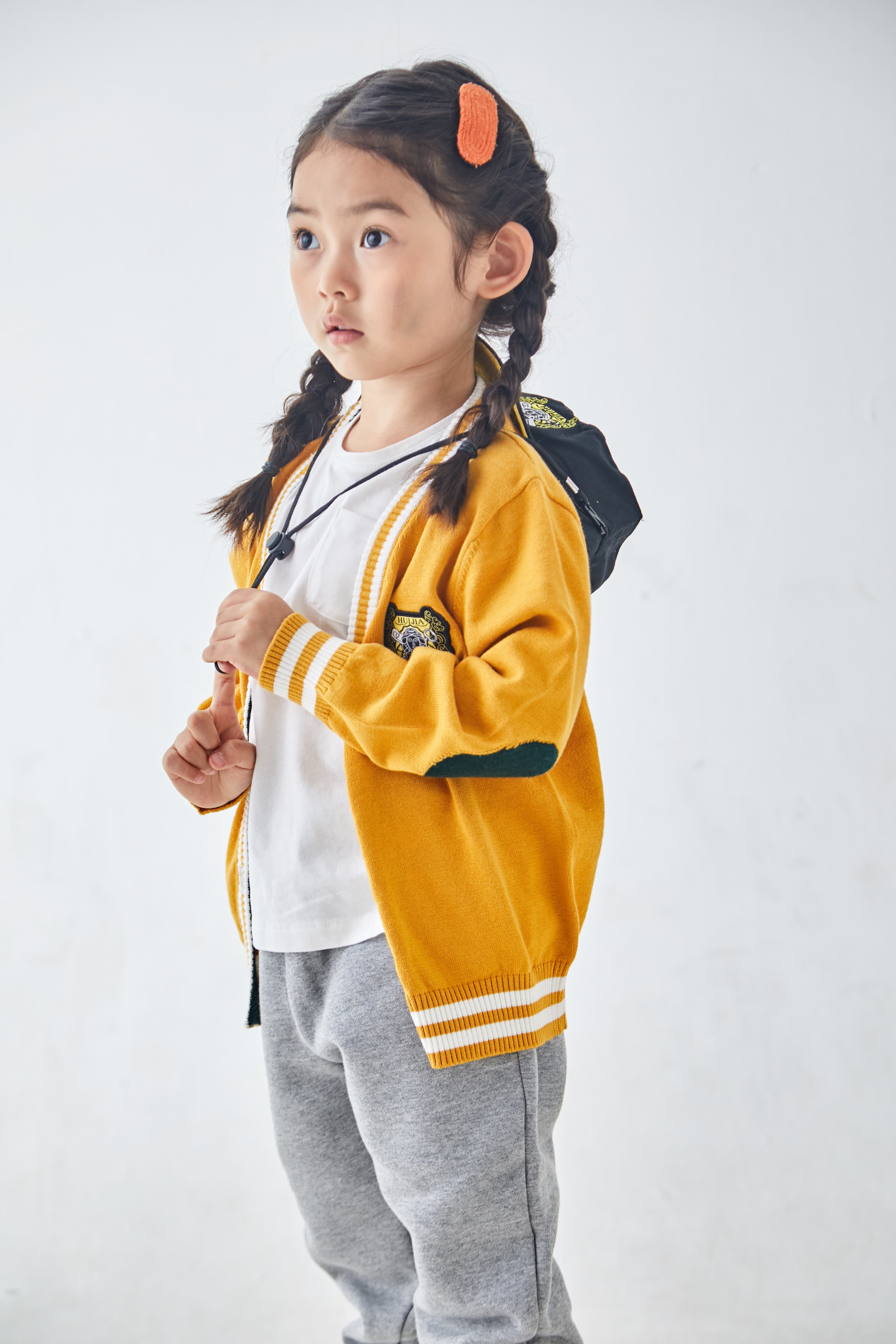 Young girl with braided hair wearing a yellow cardigan and gray pants against white background.