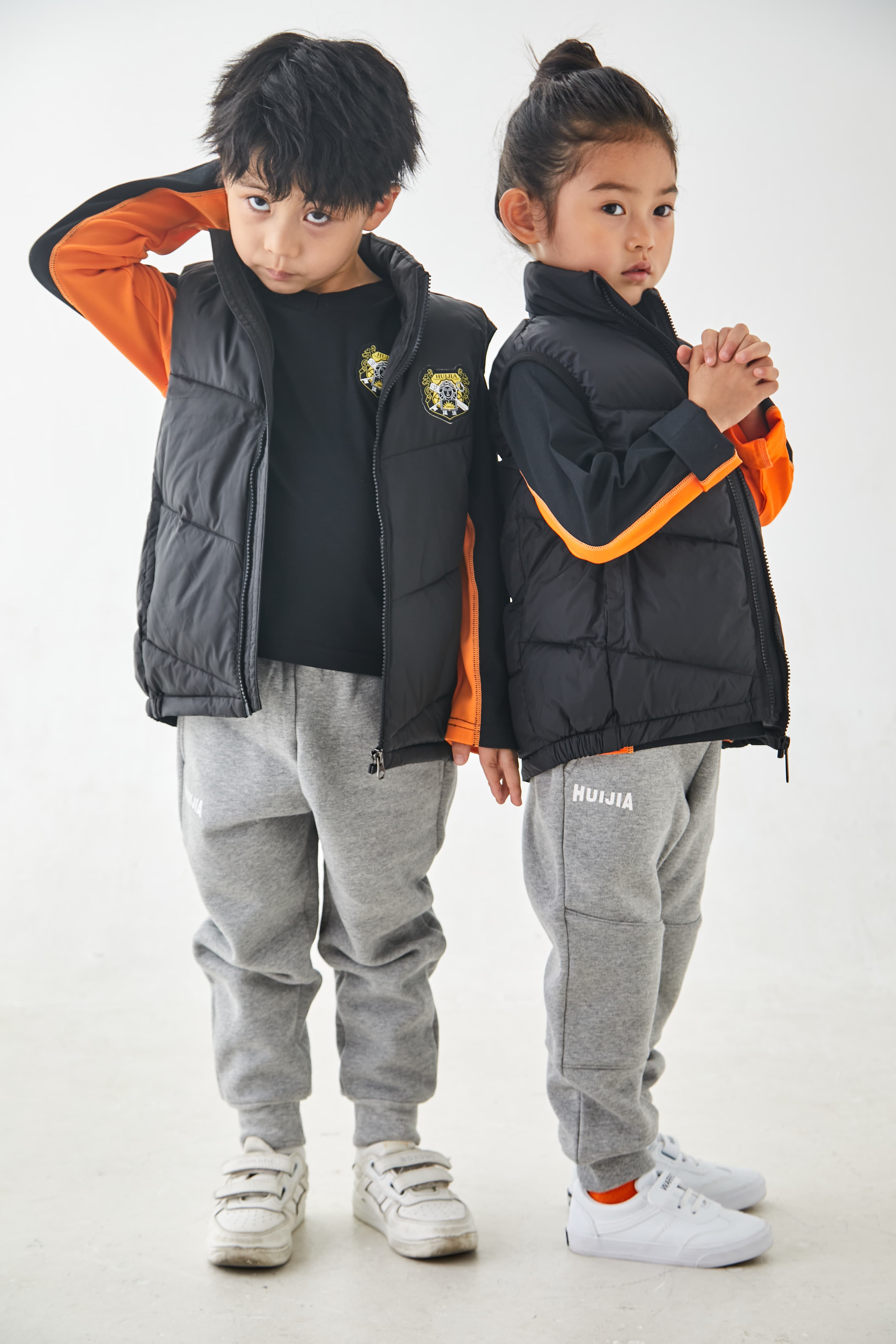 Two children posing in matching black puffer vests, orange-trimmed shirts, and grey sweatpants.